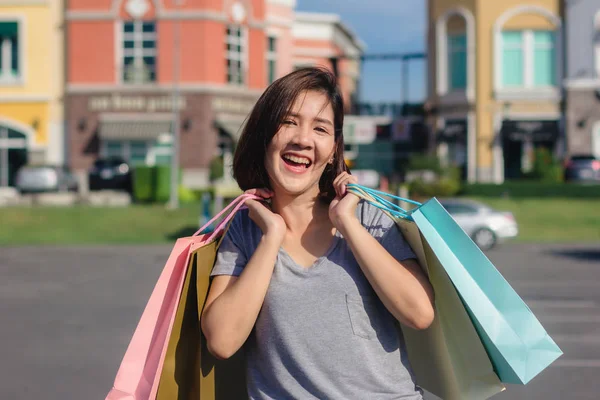 Happy young Asian woman shopping an outdoor market with background of pastel buildings and blue sky. Young asian woman smile with a colorful bag in her hand. Outdoor woman lifestyle shopping concept.