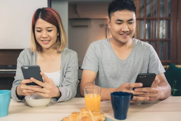 Attractive young Asian couple distracted at table with newspaper and cell phone while eating breakfast. Excited young Asian couple surprised by unbelievably good news, happy family amazed by internet.