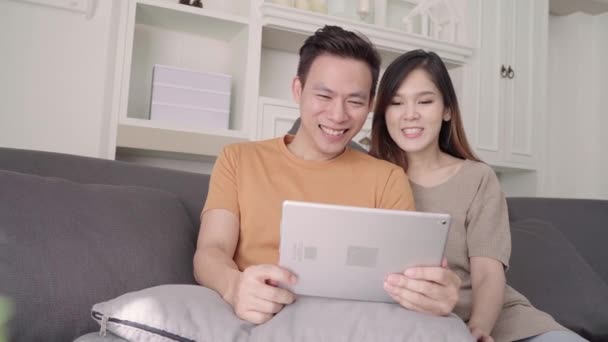 亚洲夫妇在家里客厅里用平板电脑上网购物 甜蜜的夫妇在家里放松的时候躺在沙发上享受爱情的时刻 生活方式夫妻在家里放松一下 — 图库视频影像