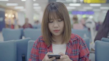 Kullanarak ve onun smartphone uçağının kalkış kapısı uluslararası havaalanında beklerken terminal Salonu sandalyede oturan süre kontrol mutlu Asyalı kadın. Kadınlar Havaalanı kavramında mutlu.