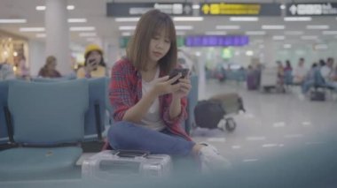 Kullanarak ve onun smartphone uçağının kalkış kapısı uluslararası havaalanında beklerken terminal Salonu sandalyede oturan süre kontrol mutlu Asyalı kadın. Kadınlar Havaalanı kavramında mutlu.