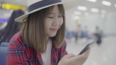 Kullanarak ve onun smartphone uçağının kalkış kapısı uluslararası havaalanında beklerken terminal Salonu sandalyede oturan süre kontrol mutlu Asyalı kadın. Kadınlar Havaalanı kavramında mutlu.