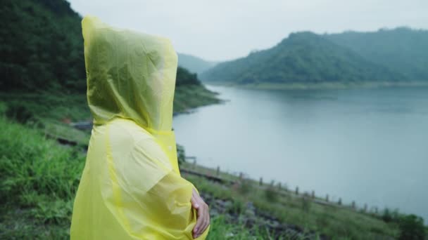 若いアジア女性感じ幸せな演奏の雨湖近く立っているレインコートを着用しながらの ライフ スタイルの女性と雨の日のリラックスをお楽しみいただけます — ストック動画