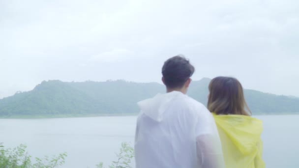 スローモーション 若いアジアの甘いカップル湖近く立っているレインコートを着て雨を再生するロマンチックな時間を使用して幸せな気持ち ライフ スタイルのカップルをお楽しみください 雨の日でリラックス — ストック動画