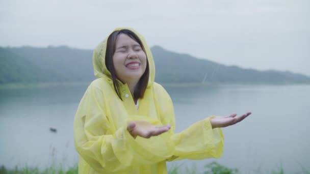 スローモーション 若いアジア女性感じ幸せ演奏湖近く立っているレインコートを着て雨 ライフ スタイルの女性と雨の日のリラックスをお楽しみいただけます — ストック動画
