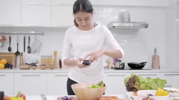 年轻的亚洲博主女性在厨房的社交媒体上使用智能手机图片帖子 在家里做沙拉 生活方式的女性放松在家里的概念 — 图库视频影像
