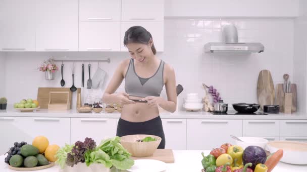キッチン 家庭でサラダを作るスポーツ服で女性の社会的なメディアのスマート フォン写真ポストを使用して若いアジアのブロガーの女性 ライフ スタイルの女性は家でくつろぐコンセプト — ストック動画