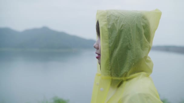 スローモーション 若いアジア女性感じ幸せ演奏湖近く立っているレインコートを着て雨 ライフ スタイルの女性と雨の日のリラックスをお楽しみいただけます — ストック動画
