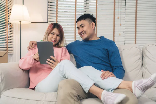 Attractive Asian sweet couple using tablet while lying on the sofa when relax in their living room at home. Husband and his wife using relax and romantic time at home concept.