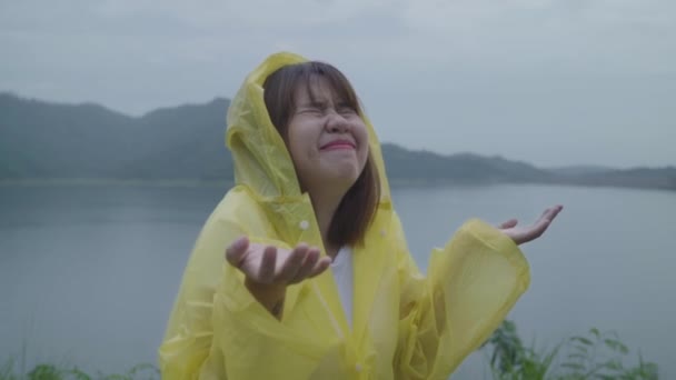 慢动作 年轻的亚洲女子穿着雨衣站在湖边玩雨 感觉很开心 生活方式的女性在雨天享受和放松 — 图库视频影像