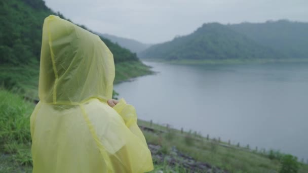 若いアジア女性感じ幸せな演奏の雨湖近く立っているレインコートを着用しながらの ライフ スタイルの女性と雨の日のリラックスをお楽しみいただけます — ストック動画