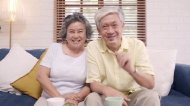 Asyalı yaşlı bir çift, torunuyla akıllı telefon görüşmesini evdeki oturma odasında kanepede yatarken yapıyor. Ev kavramındaki son sınıf ailesinin tadını çıkarıyorum. Kameraya bakan portre.