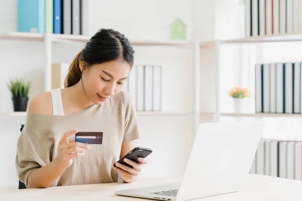 Beautiful Asian woman using smartphone buying online shopping by credit card while wear sweater sitting on desk in living room at home. Lifestyle woman at home concept.