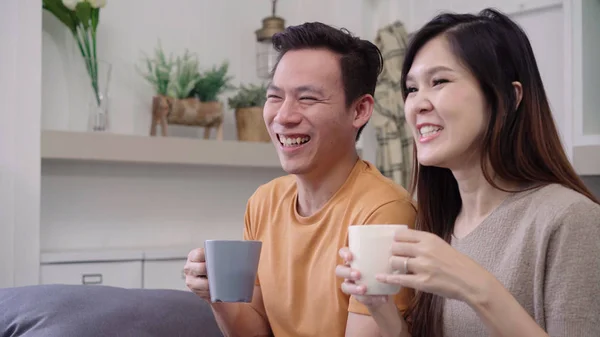 Asian couple watching TV and drinking warm cup of coffee in living room at home, sweet couple enjoy love moment while lying on the sofa when relax at home. Lifestyle couple relax at home concept.
