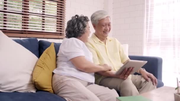 アジアの老夫婦は 自宅のリビング ルームでテレビを見ているタブレットを使用して カップルは 家でゆっくりとソファの上に横たわっている間愛の瞬間をお楽しみください ホーム コンセプトで時間ライフ スタイル シニア家族を楽しんでください — ストック動画