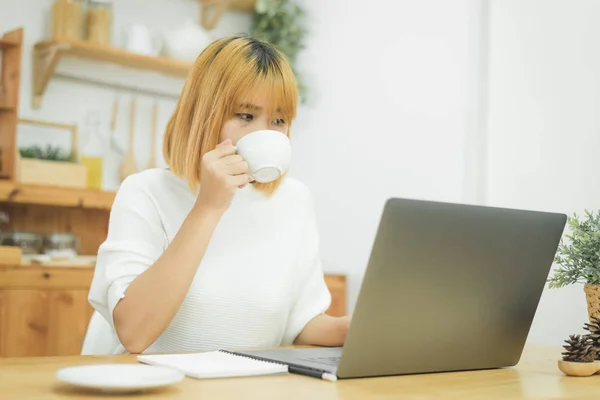 Молодая Улыбчивая Женщина Работает Ноутбуке Наслаждаясь Горячим Кофе Сидя Дома — стоковое фото