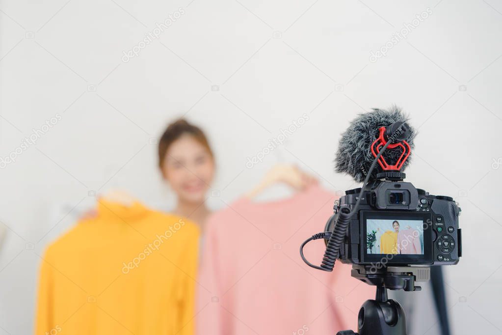 Asian fashion female blogger online influencer holding shopping bags and lots of clothes on clothes rack for recording new fashion video broadcast live video to social network by internet at home.