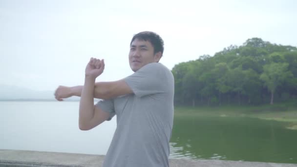 アジア ランナー男性足温暖化と湖の近くの通り 健康的な若い男性運動を実行するための準備をする彼女の腕を伸ばします ライフ スタイルに合わせて アクティブな男性運動ストリート コンセプト — ストック動画