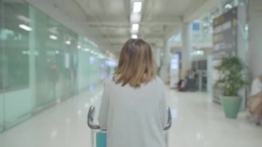 Birçok bagajlı terminal salonunda Uluslararası Havaalanı kalkış kapısında yatılı uçuşa giderken yürüme arabası veya sepeti kullanarak mutlu Asyalı kadın. Kadınlar Havaalanı kavramında mutlu.