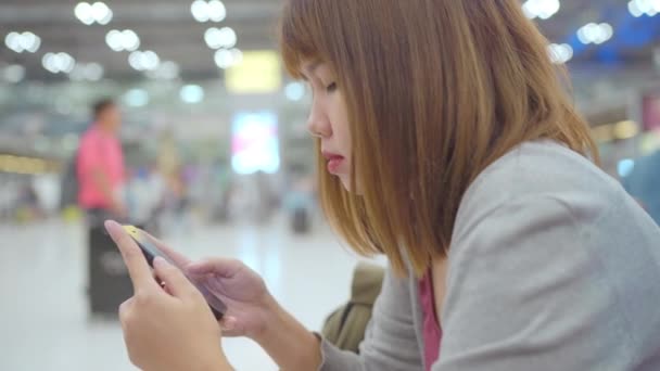 愉快的亚洲妇女使用和检查她的智能手机 而坐在候机楼的椅子上 而她的航班在国际机场的登机口等待 妇女高兴在机场概念 — 图库视频影像