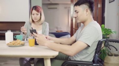 Bir tekerlekli sandalyede Mutlu tatlı Asya çift adam kahvaltı, süt, ekmek tahıl ve sabah uyandıktan sonra haber okuma cep telefonu kullanarak. Karı ve eşi birlikte yemek yiyor..