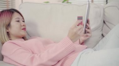 Yeni eve taşınan genç Asyalı kadın, tablet ilerletmeye devam eden güzel kadın sosyal medyayı kullanarak evde oturma odasında eve taşındıktan sonra rahatlarken kanepeye yatmış. Evde zaman yaşam tarzı kadın zevk kavramı.