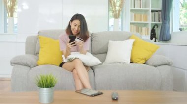 Genç gülümseyen Asyalı kadın evde oturma odasında dinlenirken kredi kartıyla online alışveriş yapıyor. Ev konseptinde Latin ve Latin kökenli kadınlar var..
