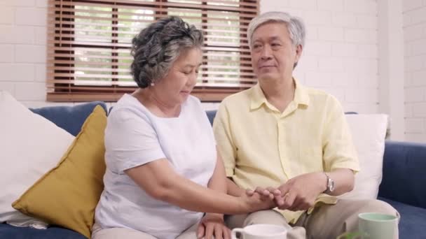居間で一緒に手をつないでいるアジアの老夫婦は 幸せな分かち合いを感じ 自宅のソファに横たわってお互いを支え合う 在宅ライフスタイルシニアファミリーコンセプト — ストック動画