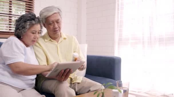 居間でタブレット検索薬情報を使用するアジアの高齢夫婦 自宅でくつろいだときにソファに横たわって一緒に時間を使うカップル 家庭での高齢者の健康コンセプト — ストック動画