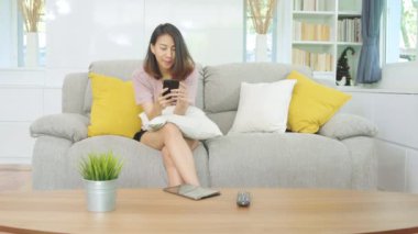 Genç Asyalı kadın akıllı telefon kullanarak sosyal medyayı kontrol ediyor. Evdeki oturma odasında dinlenirken kanepede yatarken gülümsüyor ve mutlu oluyor. Ev konseptinde Latin ve Latin kökenli kadınlar var..