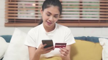 Genç gülümseyen Asyalı kadın evde oturma odasında dinlenirken kredi kartıyla online alışveriş yapıyor. Ev konseptinde Latin ve Latin kökenli kadınlar var..