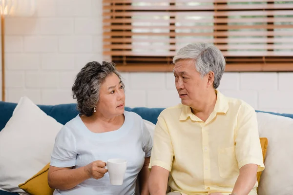 Asian elderly couple drinking warm coffee and talking together in living room at home, couple enjoy love moment while lying on sofa when relaxed at home. Lifestyle senior family at home concept. — Stock Photo, Image