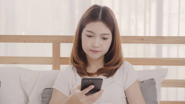 Ung asiatisk kvinne som bruker smarttelefon mens hun ligger på senga etter å ha våknet om morgenen, vakker, attraktiv japansk jente som smiler, slapper av på soverommet hjemme. Nyter tid - kvinner i hjemmet . – stockfoto