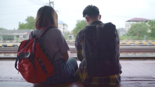 列車を待っている間にベンチに座っている旅行者アジアのバックパックカップル 若い甘いカップルの観光バックパッカーは タイでの旅を楽しみます ライフスタイルティーンカップルリラックスして旅行の概念 — ストック動画