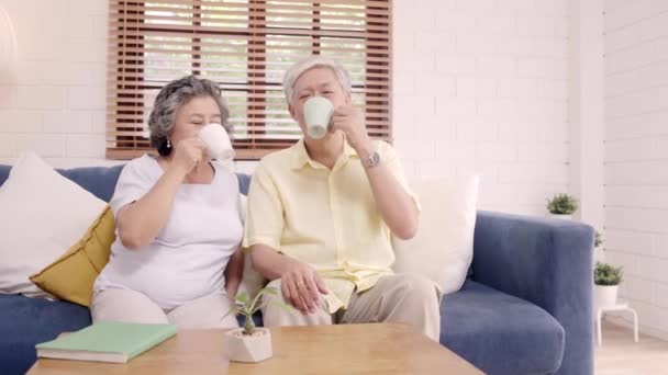 温かいコーヒーを飲み 自宅のリビングルームで一緒に話すアジアの高齢夫婦は 自宅でリラックスした時にソファに横たわりながらラブモーメントを楽しむ 在宅ライフスタイルシニアファミリーコンセプト — ストック動画
