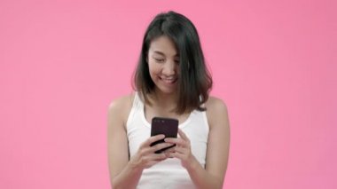 Genç Asyalı kadın pembe arka plan üzerinde rahat giyim gülümseyerek mutlu duygu sosyal medya kontrol akıllı telefon kullanarak. Mutlu gülümseyen sevimli mutlu kadın başarı sevinir.