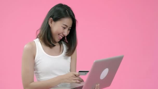 カジュアルな服を着たクレジットカードでオンラインショッピングを購入し ピンクの背景スタジオショットの上にコンピュータを見て ラップトップで使用する若いアジアの女性 幸せな笑顔愛らしい喜んで女性は成功を喜ぶ — ストック動画