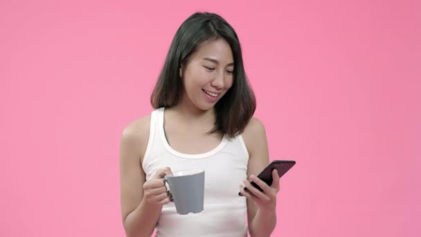 コーヒーを飲み スマートフォンを使用して ピンクの背景スタジオショットの上にカジュアルな服で幸せな感じソーシャルメディアをチェックして 若いアジアの女性 幸せな笑顔愛らしい喜んで女性は成功を喜ぶ — ストック動画