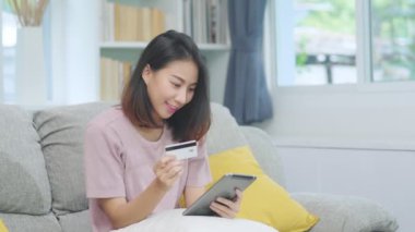 Genç gülümseyen Asyalı kadın evde oturma odasında dinlenirken kredi kartıyla online alışveriş yapıyor. Ev konseptinde Latin ve Latin kökenli kadınlar var..