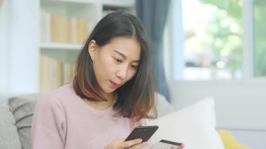 Genç gülümseyen Asyalı kadın evde oturma odasında dinlenirken kredi kartıyla online alışveriş yapıyor. Ev konseptinde Latin ve Latin kökenli kadınlar var..