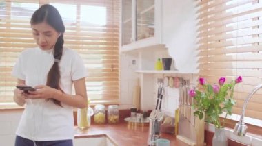Genç Asyalı genç kadın akıllı telefon müzik dinleme ve evde mutfakta sosyal kontrol kullanarak sıcak kahve içme. Yaşam tarzı kadın evde kavramı sabah dinlenmek.