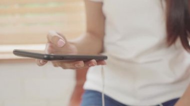 Genç Asyalı genç kadın akıllı telefon müzik dinleme ve evde mutfakta sosyal kontrol kullanarak sıcak kahve içme. Yaşam tarzı kadın evde kavramı sabah dinlenmek.