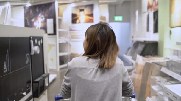 年轻的亚洲妇女骑着购物车在仓库里挑选新家具 在百货公司的货架前 年轻的女性感到快乐 女性购买新家具概念 — 图库视频影像