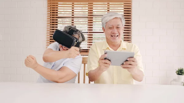 Asian elderly couple using tablet and virtual reality simulator playing games in living room, couple feeling happy using time together lying on table at home. Lifestyle Senior family at home concept.