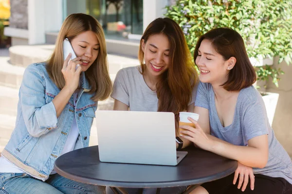 Mujeres jóvenes asiáticas que trabajan en el ordenador portátil usando y mirando teléfono inteligente y beber café mientras están sentadas en la cafetería. Estilo de vida de las mujeres de comunicación y trabajo en concepto de cafetería . — Foto de Stock
