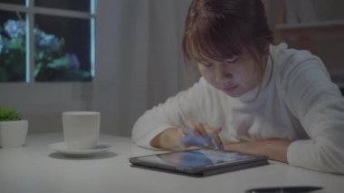 Genç asyalı kadın evde oturma odasında gece masada dinlenirken sosyal medya kontrol tablet kullanarak geç çalışıyor. Evde vakit geçirme konsepti.