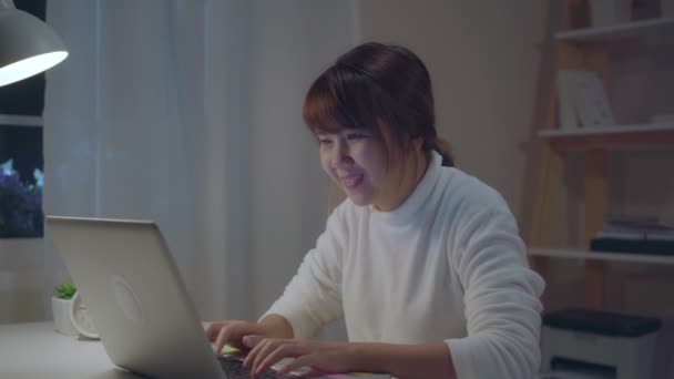 年轻的亚洲妇女工作到很晚 在家里客厅的办公桌上使用笔记本电脑 亚洲女商人成功庆祝 晚上在家里的办公室举起手臂 享受在家的时间概念 — 图库视频影像
