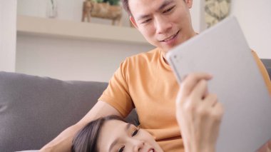 Evdeki oturma odasında internet alışverişi için tablet kullanan Asyalı çift, evde dinlenirken kanepede uzanırken aşk anının tadını çıkarıyor. Yaşam tarzı çifti ev kavramında rahatlar..