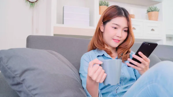 Portrett av en vakker, smilende asiatisk kvinne som bruker smarttelefon med en varm kopp kaffe eller te mens hun ligger på sofaen når hun slapper av i stua. Livsstilsvenninner hjemme . – stockfoto