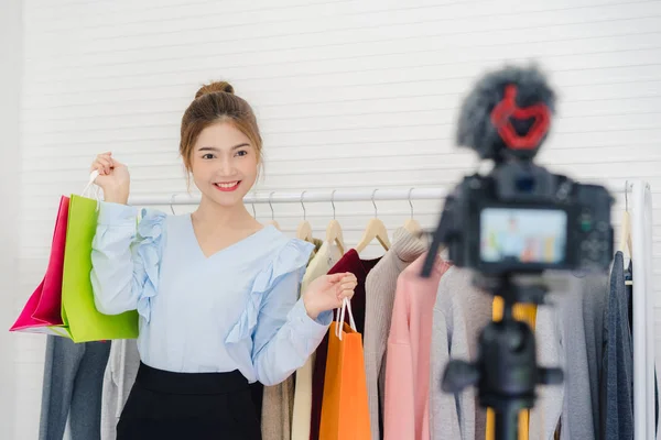 Asiatisk mode kvinnliga bloggare online påverkare hålla shoppingkassar och massor av kläder på klädhängare för inspelning av nya mode video sändning live video till sociala nätverk av internet hemma. — Stockfoto